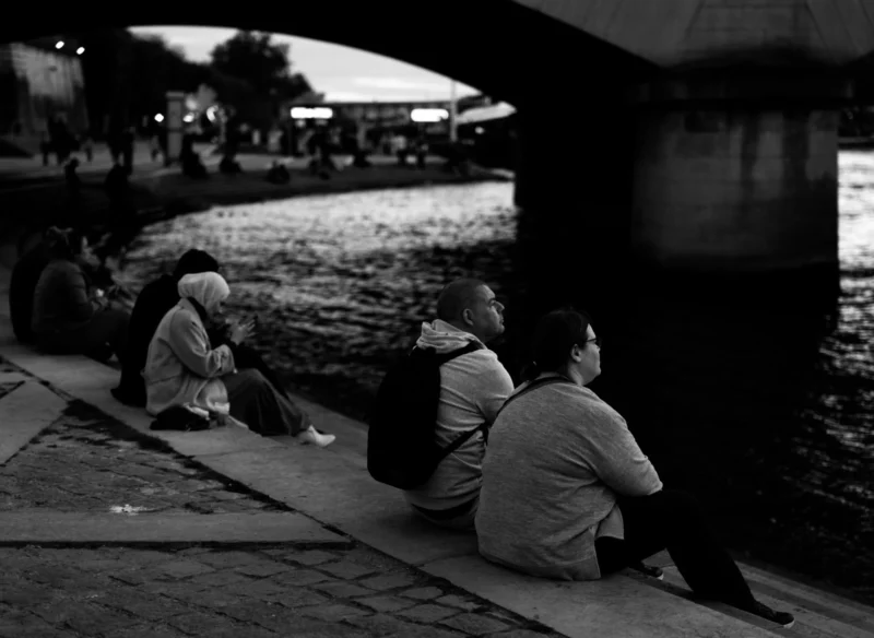 Bao · 2369 x 1730 Cette photographie en noir et blanc met en scène trois personnes assises sur une berge, près d’un canal. Le sujet principal est ce groupe en conversation, créant une ambiance intimiste et contemplative.La composition est équilibrée, avec un cadrage serré qui isole les personnages du reste de l’environnement. La lumière douce, probablement crépusculaire, adoucit les contours et ajoute une touche mélancolique. Les couleurs dominantes, bien que limitées au noir et blanc, suggèrent des tons gris et blancs, renforçant l’aspect sobre et introspectif.Le style est documentaire et réaliste, évoquant une scène du quotidien. L’ambiance est calme et légèrement nostalgique.