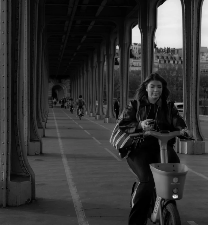 Bao · 1781 x 1923 Cette photographie en noir et blanc met en scène une femme comme sujet principal, assise sur un vélo sous une passerelle. La composition est symétrique, avec une perspective guidée par les colonnes et les lignes architecturales.Le cadrage est serré sur la personne, mais inclut aussi l’environnement urbain en arrière-plan. La lumière est douce, sans ombres marquées, créant une ambiance calme et mélancolique. Les couleurs dominantes, bien que monochromes, jouent sur les contrastes de gris et de noir, renforçant l’aspect intemporel et artistique du cliché. Le style est documentaire et esthétique, évoquant une atmosphère contemplative et urbaine.