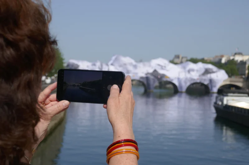 Bao · 5472 x 3648 Cette photographie montre une personne prenant une photo avec un smartphone. Le sujet principal est le reflet des arches blanches sur l’eau, probablement un pont ou des sculptures. La composition est centrée sur le téléphone, encadrant partiellement le paysage.Le cadrage est serré sur les mains et le smartphone, avec un arrière-plan flou mais identifiable. La lumière est naturelle et claire, suggérant un jour ensoleillé. Les couleurs dominantes sont les tons blancs des arches, le bleu du ciel et de l’eau, et les teintes chaudes des bracelets. L’ambiance est paisible et artistique, mettant en valeur la beauté architecturale et la sérénité du lieu.