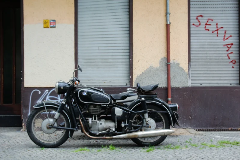Cédric · 23/05/2017 · 2048 x 1365 Cette photographie met en scène une **moto vintage** comme sujet principal, probablement des années 1950. Le **cadrage** est serré, centré sur le véhicule, avec un arrière-plan minimaliste : un mur tagué ("SEX SHOP") et une porte.La **composition** est équilibrée, avec une légère perspective en contre-plongée. La **lumière** semble naturelle, douce, avec des ombres marquées, suggérant un éclairage matinal ou vespertin. Les **couleurs dominantes** sont les tons gris métallisés de la moto, contrastant avec le vert des graffitis et le beige du mur.L’**ambiance** est à la fois **nostalgique** et **urbaine**, mêlant charme rétro et réalisme de la ville.