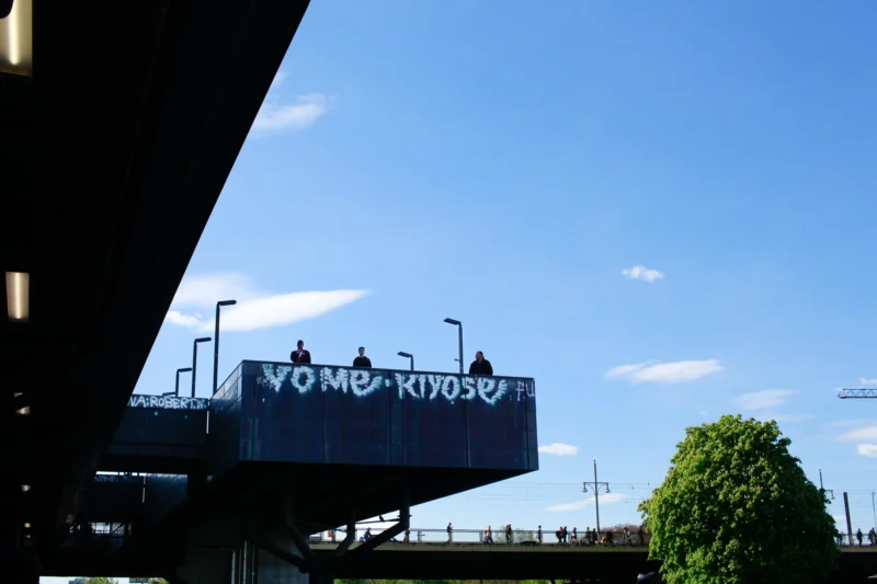 Cédric · 30/04/2026 · 2048 x 1365 Cette photographie montre un panneau publicitaire ou une structure sur un pont avec l'inscription "VO MERCIOS" en lettres stylisées. Le sujet principal est ce message, accompagné de silhouettes humaines.La composition inclut un cadrage en contre-plongée sous une structure métallique, mettant en valeur le ciel bleu et quelques nuages. La lumière est naturelle, avec un éclairage doux, créant des contrastes nets. Les couleurs dominantes sont les bleus du ciel et les tons gris métalliques du pont. Le style est épuré et urbain, évoquant une ambiance moderne et légèrement mystérieuse.