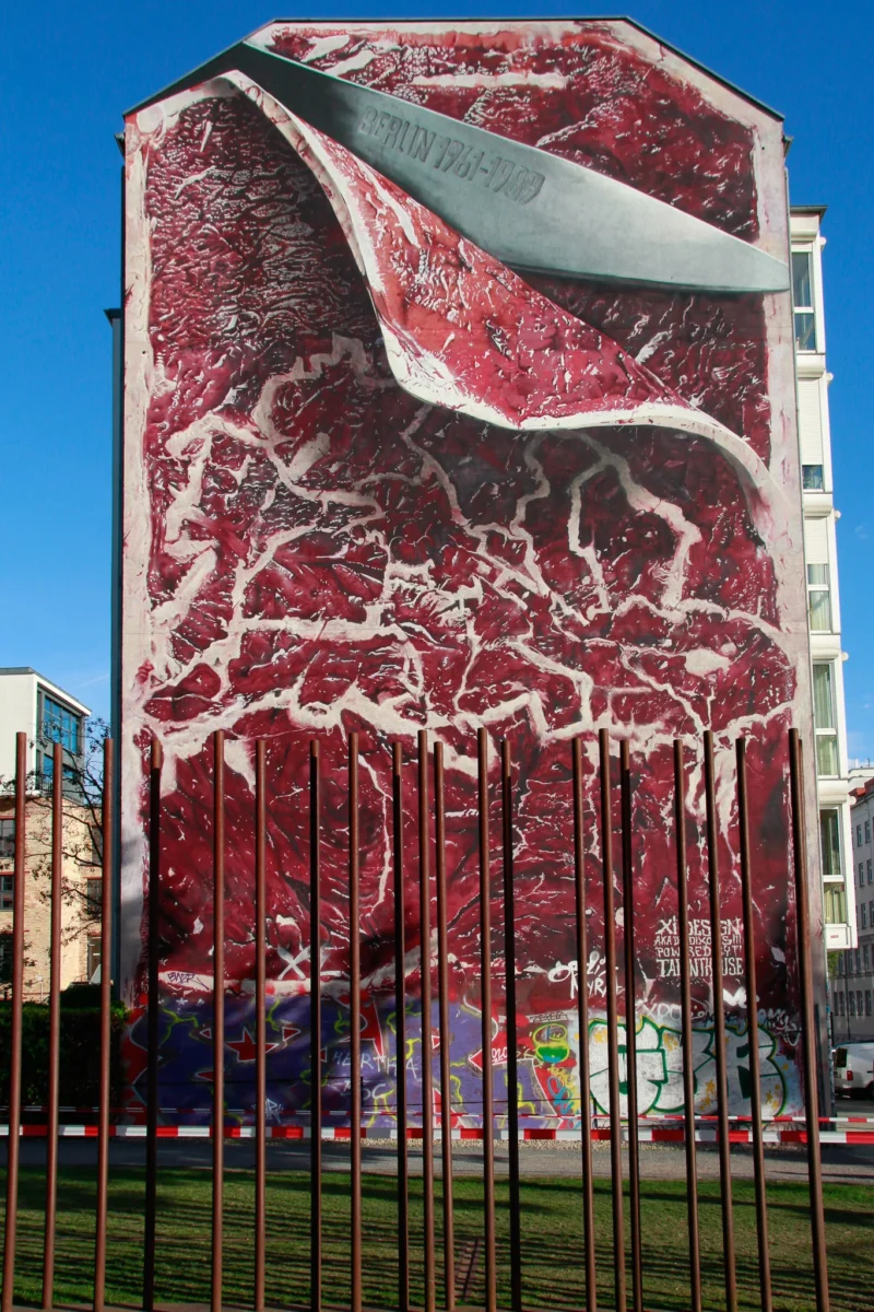 Cédric · 30/04/2026 · 1365 x 2048 Cette photographie montre une façade d’immeuble recouverte d’un mural représentant une tranche de viande crue, avec des vaisseaux sanguins rouges et blancs. Le sujet principal est cette œuvre artistique frappante.La composition est verticale, avec un cadrage serré sur le mural. Les couleurs dominantes sont le rouge sang et le blanc, créant un contraste saisissant. La lumière naturelle éclaire la scène, accentuant les détails. Le style est réaliste et provocateur, évoquant une ambiance à la fois artistique et dérangeante, mêlant beauté et brutalité.