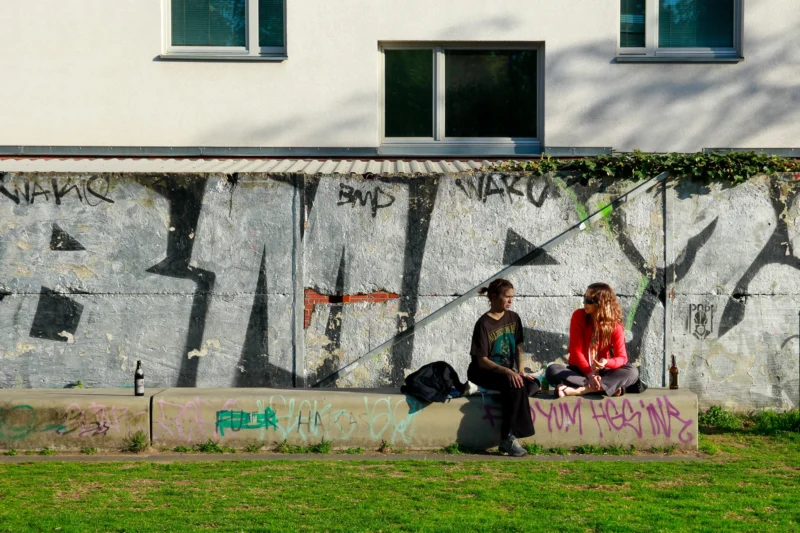Cédric · 30/04/2026 · 2048 x 1365 Cette photographie met en scène deux personnes assises sur un mur tagué, discutant. Le sujet principal est leur interaction, dans une ambiance détendue.La composition repose sur un cadrage serré, mettant l’accent sur les personnages et le graffiti coloré. La lumière naturelle, douce et légèrement contrastée, éclaire la scène, créant des ombres subtiles.Les couleurs dominantes sont les tons gris du mur, rehaussés par des graffitis vifs (rouge, orange, noir). Le style est documentaire et réaliste, évoquant une ambiance urbaine et spontanée.