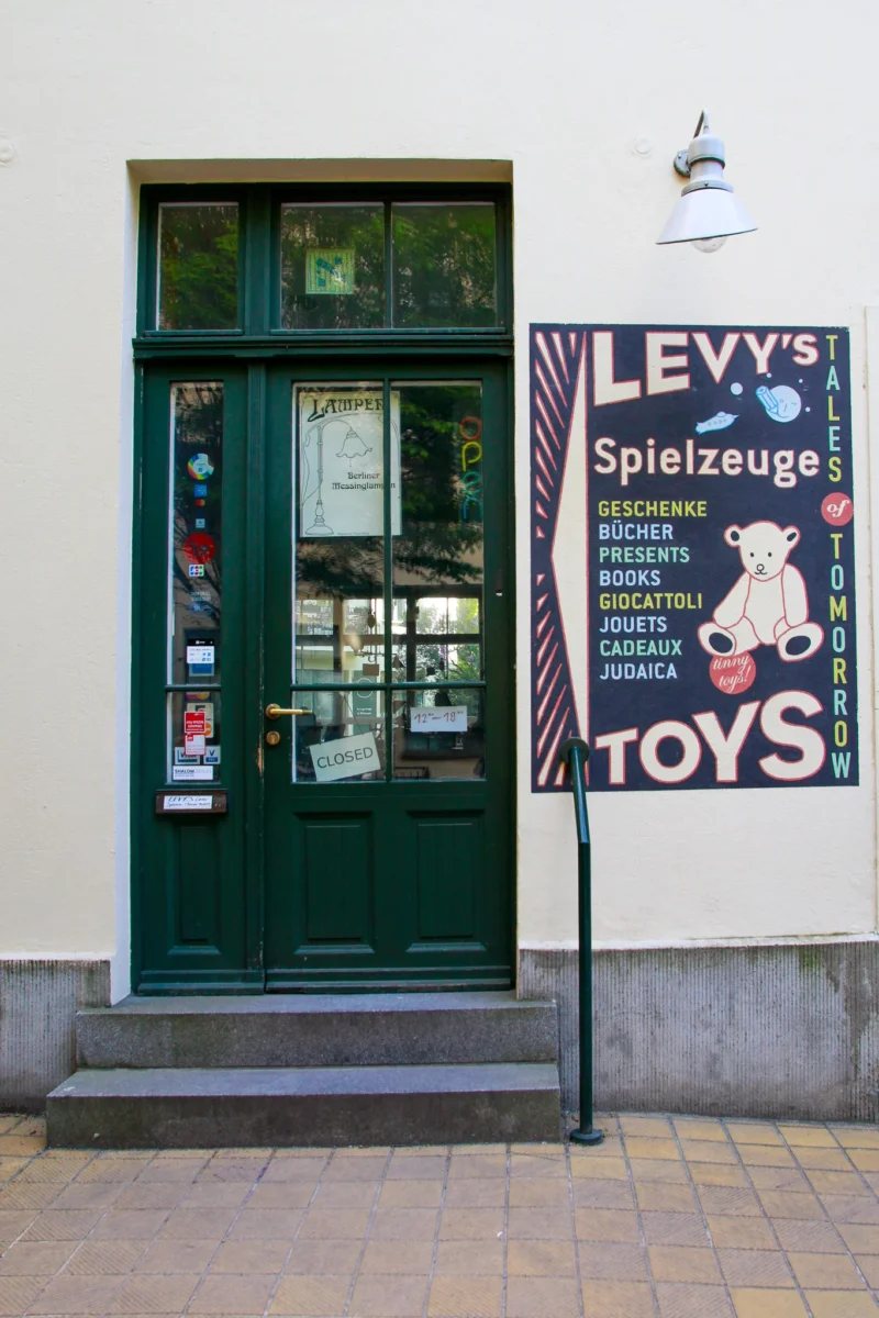 Cédric · 01/05/2026 · 1365 x 2048 Cette photographie montre une boutique nommée **Levy’s Spielzeuge**, spécialisée dans les jouets. Le sujet principal est l’entrée verte avec une vitrine et un panneau publicitaire coloré en noir et blanc, mettant en avant des jouets et des peluches.La composition est symétrique, centrée sur la porte. Le cadrage est serré, capturant uniquement la façade. La lumière est naturelle et douce, éclairant bien la scène. Les couleurs dominantes sont le vert de la porte, le noir et blanc du panneau, et des touches de rouge et bleu.Le style est épuré et commercial, évoquant une ambiance chaleureuse et conviviale, typique d’une boutique de jouets.
