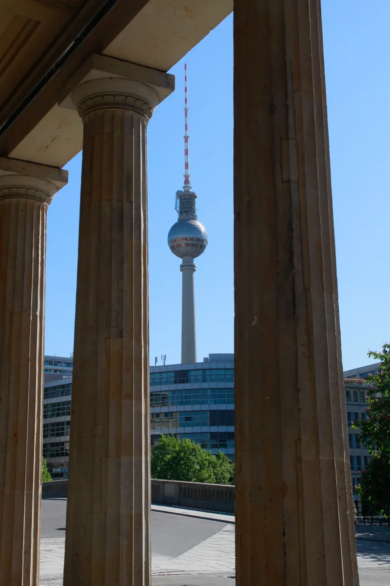 Cédric · 01/05/2026 · 1365 x 2048 Cette photographie met en scène la **Tour de télévision de Berlin** (Fernsehturm) comme sujet principal. Le cadrage est effectué à travers deux **colonnes classiques**, créant un effet de perspective et de profondeur.La lumière est naturelle et claire, typique d’un jour ensoleillé. Les couleurs dominantes sont les **tons gris-beige des colonnes**, contrastant avec le **bleu du ciel** et le **vert des arbres** en arrière-plan. Le style est épuré et architectural, évoquant une ambiance à la fois **classique et moderne**. L’image dégage une sensation de sérénité et de grandeur urbaine.