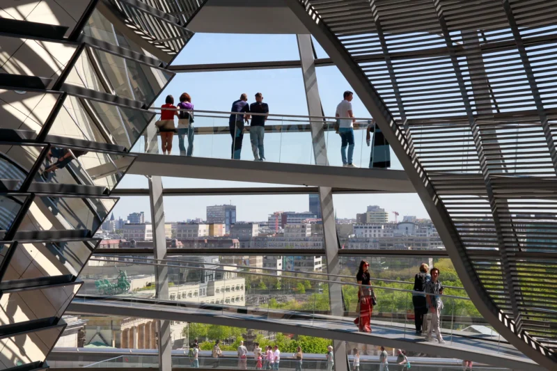 Cédric · 01/05/2026 · 2048 x 1365 Cette photographie montre l'intérieur du **Dôme de verre du Reichstag** à Berlin. Le sujet principal est l’architecture moderne avec ses structures géométriques en verre et métal. La composition met en valeur les visiteurs sur une passerelle transparente, offrant une vue panoramique sur la ville.Le cadrage est large, capturant à la fois les détails architecturaux et le paysage urbain. La lumière est naturelle et abondante, traversant les vitres. Les couleurs dominantes sont les tons froids du verre et du métal, contrastant avec les teintes chaudes de la ville en arrière-plan.L’ambiance est à la fois futuriste et transparente, symbolisant l’ouverture et la modernité.