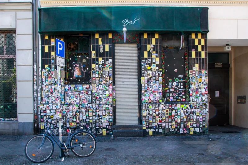 Cédric · 02/05/2026 · 2048 x 1365 Cette photographie montre une boutique dont les portes vitrées sont recouvertes de nombreux cadenas colorés, typiques des "love locks". Le sujet principal est cette accumulation de cadenas symbolisant l’amour.La composition est symétrique, avec un cadrage serré sur les portes. Un vélo est partiellement visible à gauche, ajoutant un élément urbain. La lumière semble naturelle, avec des ombres douces. Les couleurs dominantes sont vives et variées grâce aux cadenas multicolores.Le style est documentaire et urbain, évoquant une ambiance à la fois romantique et mélancolique, liée aux serments d’amour et à leur abandon.