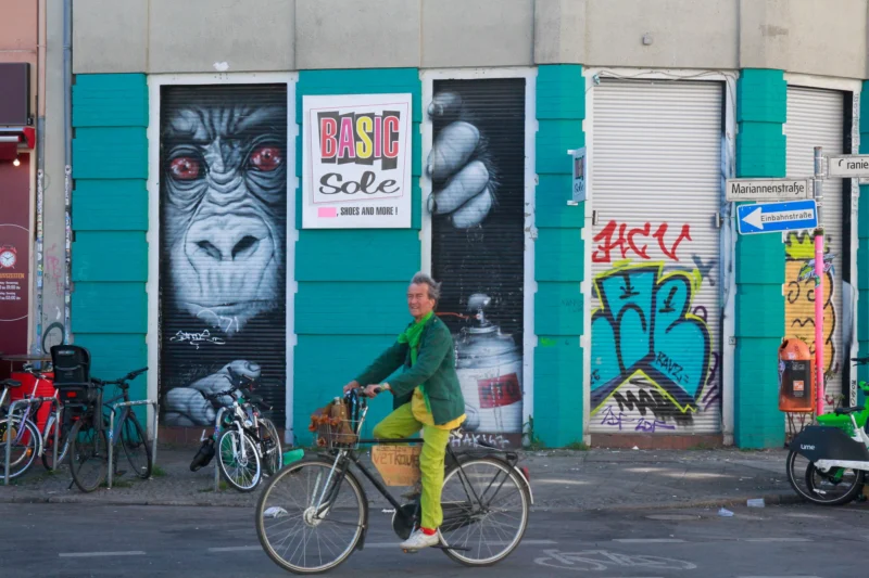 Cédric · 02/05/2026 · 2048 x 1365 Cette photographie met en scène un cycliste vêtu d’un pantalon jaune vif, sujet principal. Le cadrage est serré sur lui, avec un arrière-plan urbain coloré : graffitis, affiches et portes closes.La composition joue sur les contrastes entre le mouvement du cycliste et l'immobilité des éléments architecturaux. La lumière semble naturelle, probablement en journée, éclairant les teintes vives.Les couleurs dominantes sont les tons verts, jaunes et bleus, renforcés par les graffitis. Le style est réaliste et spontané, capturant une ambiance urbaine dynamique et artistique.