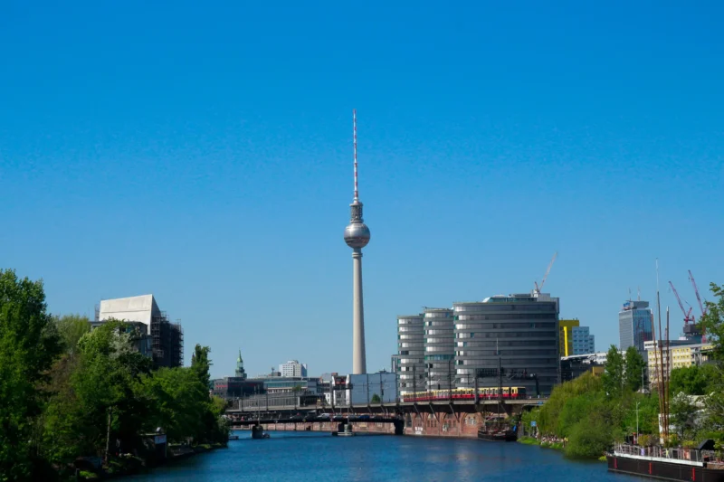 Cédric · 02/05/2026 · 2048 x 1365 Cette photographie met en scène la **Tour de télévision de Berlin**, sujet principal, se détachant sur un ciel dégagé. La composition montre un cadre urbain avec des bâtiments modernes et un pont en arrière-plan, encadré par des arbres en bas.Le cadrage est large, capturant une vue panoramique. La lumière est naturelle, vive et claire, suggérant un jour ensoleillé. Les couleurs dominantes sont les **bleus du ciel**, les **verts des arbres**, et les **tons gris-beige des bâtiments**.Le style est photographique réaliste, avec une ambiance sereine et moderne, reflétant une ville dynamique et bien organisée.