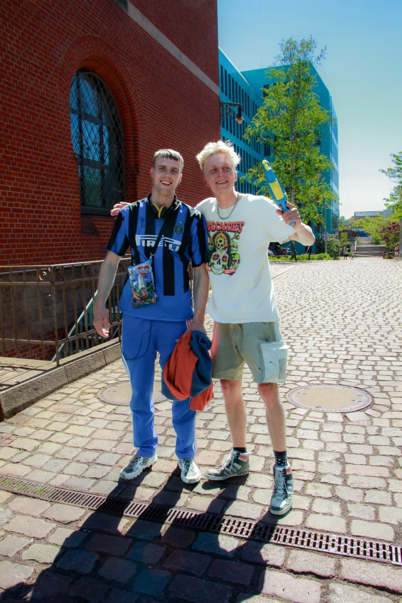 Cédric · 02/05/2026 · 1365 x 2048 Cette photographie montre deux personnes souriantes posant devant un bâtiment. Le sujet principal est ces deux individus, l’un avec un sac à dos et un t-shirt coloré, l’autre en t-shirt blanc et short.La composition est symétrique, avec un cadrage serré sur les deux personnes. La lumière est naturelle et éclatante, suggérant un jour ensoleillé. Les couleurs dominantes sont les teintes vives des vêtements et le rouge du bâtiment en arrière-plan.Le style est simple et spontané, évoquant une ambiance détendue et joyeuse, typique d’un voyage ou d’une excursion.