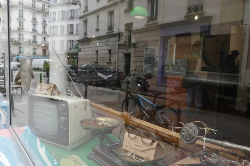 Bao · 6192 x 4128 Cette photographie montre une vitrine remplie d’objets vintage. Le sujet principal est un vieux téléviseur noir et blanc posé sur une étagère, entouré d’autres articles rétro comme des bicyclettes et des accessoires anciens.La composition est désordonnée mais harmonieuse, avec un cadrage serré sur les objets. La lumière est naturelle, tamisée, créant des reflets sur la vitrine. Les couleurs dominantes sont neutres (gris, beige) avec quelques touches de vert et bleu.Le style évoque un dépaysement nostalgique, tandis que l’ambiance, à la fois rétro et urbaine, donne une impression de curiosité et de charme vintage.