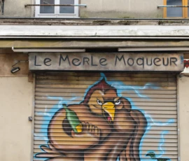 Fahad · 14/05/2026 · 2000 x 3000 Cette photographie montre une enseigne de magasin nommée **« Le Merle Moqueur »** avec un store métallique décoré d’un graffiti coloré. Le sujet principal est un octopus stylisé, entouré de fruits et de motifs psychédéliques.La composition est équilibrée, centrée sur le store. Le cadrage est serré, mettant en valeur les détails du graffiti et de l’écriture. La lumière semble naturelle, probablement en journée, avec des ombres douces.Les couleurs dominantes sont vives : bleu, orange, jaune et rouge. Le style est urbain et artistique, mêlant street art et écriture manuscrite. L’ambiance est dynamique et créative, reflétant une touche de rébellion et d’originalité.