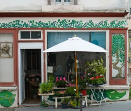 Fahad · 14/05/2026 · 3000 x 2000 Cette photographie montre une boutique fleurie avec une façade colorée. Le sujet principal est l’entrée de la boutique, décorée de plantes et de fleurs.La composition met en avant un cadrage serré sur la vitrine et l’auvent blanc. La lumière naturelle éclaire la scène, créant des ombres douces. Les couleurs dominantes sont les tons pastel (rose, vert menthe) et le blanc, renforçant une ambiance chaleureuse et accueillante. Le style est simple et pittoresque, évoquant un charme artisanal et rustique. L’ambiance est conviviale et végétale, typique d’un petit commerce de fleurs.