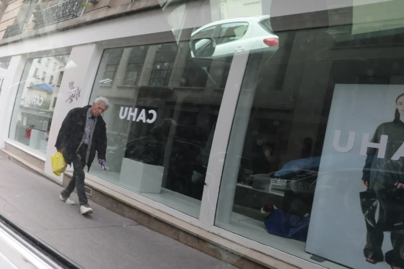 Bao · 6192 x 4128 Cette photographie montre un homme marchant devant une vitrine de magasin avec l’enseigne "CAHU". Le sujet principal est cet homme, vêtu d’un manteau sombre et portant un sac jaune.La composition est simple, avec un cadrage serré sur la vitrine et le piéton. La lumière semble naturelle, légèrement tamisée, créant des ombres douces. Les couleurs dominantes sont les tons gris et neutres, avec des touches de jaune et de blanc.L’ambiance est urbaine et quotidienne, reflétant une scène banale en ville. Le style est réaliste et documentaire.