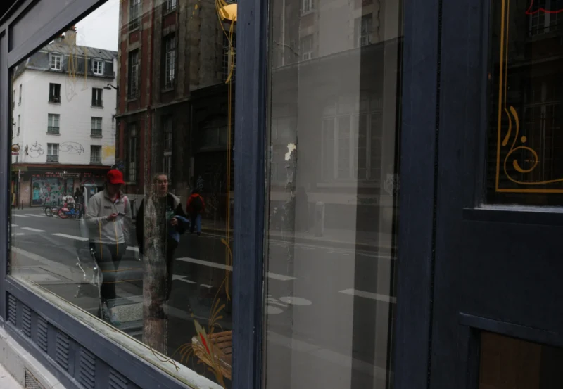 Bao · 5619 x 3880 Cette photographie capture des reflets dans une vitrine. Le sujet principal est la représentation inversée de deux personnes et de leur environnement urbain, visible à travers la vitre.La composition utilise un cadrage serré sur la vitrine, créant un effet miroir. La lumière est naturelle mais tamisée, donnant une ambiance sobre et légèrement grise. Les couleurs dominantes sont des tons neutres : gris, beige et quelques touches de rouge et noir.Le style est réaliste et documentaire, évoquant une ambiance urbaine calme et introspective.