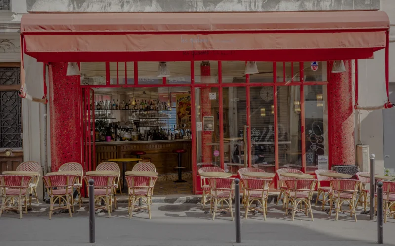 Nathalie C H · 02/05/2026 · 2000 x 1248 Cette photographie montre une terrasse de café urbaine. Le sujet principal est l’espace extérieur avec ses tables et chaises en métal, disposées sous un auvent rouge. La composition est symétrique, avec des rangées de tables alignées.Le cadrage est serré, centré sur la terrasse. La lumière semble naturelle, douce, probablement en journée. Les couleurs dominantes sont le rouge vif de l’auvent et des chaises, contrastant avec le beige des murs et le gris de la chaussée.Le style est épuré et moderne, évoquant une ambiance chaleureuse et conviviale, typique d’un café parisien.
