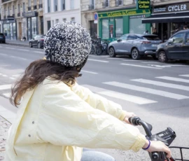Nathalie C H · 16/05/2026 · 1294 x 1188 Cette photographie montre une personne à vélo, vue de dos, comme sujet principal. Elle porte un manteau jaune pâle et un chapeau à motifs noirs et blancs.La composition met en valeur la rue urbaine avec des bâtiments, des voitures et des enseignes. Le cadrage est serré sur le cycliste, avec une profondeur de champ qui inclut l’arrière-plan flou.La lumière semble naturelle, probablement en journée. Les couleurs dominantes sont les tons pastel (jaune, gris) et les contrastes urbains (noir, blanc).Le style est documentaire et réaliste, capturant une scène de vie quotidienne dans une ambiance calme et moderne.