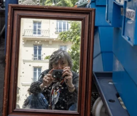 Nathalie C H · 16/05/2026 · 1159 x 1213 Cette photographie montre une personne prenant une photo dans un miroir accroché à l’extérieur. Le sujet principal est le reflet de la personne avec son appareil photo.La composition joue sur la symétrie et la réflexion, avec un cadrage serré sur le miroir. L’arrière-plan révèle des bâtiments et une ambiance urbaine. La lumière est naturelle, probablement en journée, avec des ombres douces.Les couleurs dominantes sont les tons neutres (beige, gris) des bâtiments, contrastés par le bleu vif du conteneur à côté. Le style est documentaire et spontané, créant une ambiance à la fois intime et urbaine.