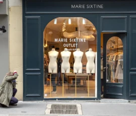 Nathalie C H · 16/05/2026 · 1099 x 798 Cette photographie montre la façade d’un outlet **Marie Sixtine**. Le sujet principal est la vitrine élégante, mettant en scène des mannequins en costumes élégants. La composition est symétrique, avec une entrée centrale et des miroirs latéraux.Le cadrage est serré sur la boutique, avec une personne assise à gauche, ajoutant une touche urbaine. La lumière est naturelle et douce, éclairant la vitrine. Les couleurs dominantes sont les tons **bleu foncé** (mur) et **beige/blanc** (vitrine et mannequins), créant une ambiance chic et sophistiquée. Le style est épuré et luxueux, évoquant le raffinement.