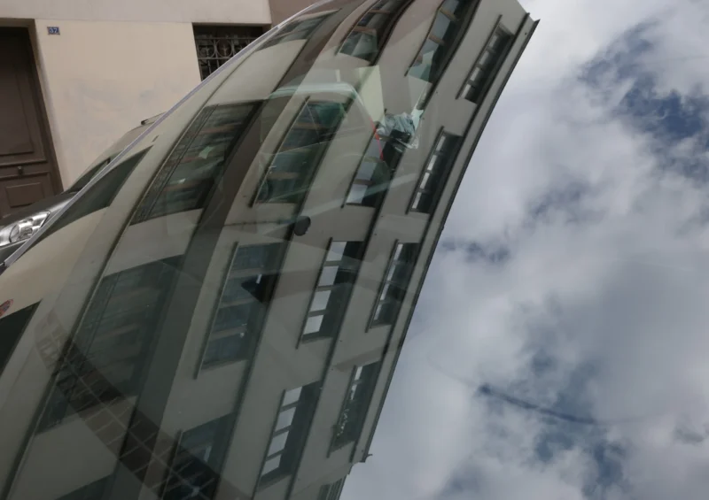 Bao · 5843 x 4128 Cette photographie met en scène un **bâtiment moderne aux lignes géométriques**, avec des **vitres réfléchissantes** et une structure métallique. Le **sujet principal** est l’architecture futuriste, vue de bas en haut, accentuant sa hauteur.Le **cadrage serré** et l’**angle incliné** créent une perspective dynamique. La **lumière naturelle**, probablement en journée, éclaire les surfaces vitrées, générant des **reflets et contrastes**. Les **couleurs dominantes** sont les tons gris (bâtiment, nuages) et bleu pâle (ciel).Le style est **urbain et contemporain**, évoquant une **ambiance technologique et minimaliste**, presque abstraite par les jeux de lumière.