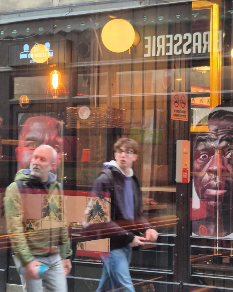 Bao · 3000 x 3752 Cette photographie montre deux hommes marchant devant une vitrine reflétante. Le sujet principal semble être la réflexion et les silhouettes floues à l'intérieur du magasin.La composition utilise le reflet pour créer un effet visuel intéressant. Le cadrage est serré sur les passants et la vitrine. La lumière est douce, probablement artificielle, avec des teintes chaudes.Les couleurs dominantes sont des tons neutres (gris, beige) avec des éclats de rouge et de bleu. Le style est urbain et spontané, évoquant une ambiance nocturne animée dans une ville.