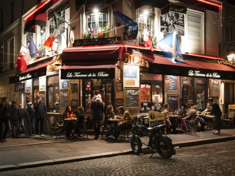 Jean-Philippe · 02/12/2025 · 13409 x 10057 Cette photographie capture une scène nocturne animée devant un restaurant parisien, "Le Tourneur de Rillettes". Le sujet principal est l'extérieur du restaurant, avec ses tables occupées et ses clients en terrasse.La composition montre une rue pavée, des chaises et tables disposées en désordre, et des personnes socialisant. Le cadrage est large, incluant une partie de la rue et des passants.La lumière est douce et artificielle, provenant des enseignes et des lampes. Les couleurs dominantes sont les tons chauds (rouge, orange) et les éclairages jaunes, créant une ambiance chaleureuse et conviviale. Le style est réaliste et documentaire.