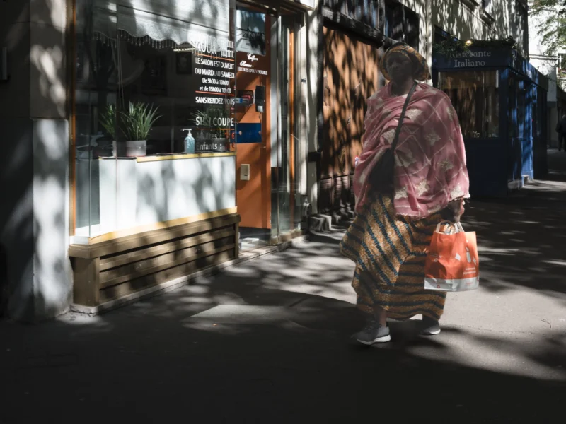 Jean-Philippe · 14/04/2026 · 6167 x 4625 Cette photographie met en scène une personne vêtue d’un long voile coloré, marchant devant une boutique artisanale. Le sujet principal est flou, attirant l’attention sur son mouvement et sa silhouette.La composition joue sur les contrastes entre lumière et ombre, avec un cadrage serré sur l’entrée de la boutique. Les jeux de lumière naturelle créent des ombres marquées, ajoutant une ambiance dramatique.Les couleurs dominantes sont les tons chauds (orange, marron) et les teintes pastel du voile. Le style est documentaire et artistique, évoquant une atmosphère authentique et contemplative, presque poétique.