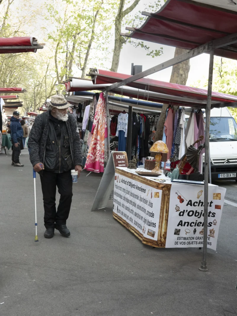 Jean-Philippe · 14/04/2026 · 3910 x 5213 Cette photographie montre un marché de rue. Le sujet principal est un homme âgé, appuyé sur un déambulateur, marchant devant des étals.La composition met en avant les stands couverts de tissus colorés et des panneaux publicitaires. Le cadrage est serré sur les étals et l’homme, avec une profondeur de champ moyenne. La lumière semble naturelle, un peu diffuse, typique d’un jour couvert.Les couleurs dominantes sont les tons pastel des tissus et les teintes neutres des structures. L’ambiance est celle d’un marché traditionnel, animé mais paisible. Le style est documentaire, captant une scène de vie quotidienne.