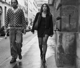 Jean-Yves · 15/05/2026 · 2935 x 3000 Cette photographie en noir et blanc met en scène deux personnes marchant sur un trottoir urbain. Le sujet principal est ce couple, dont les styles vestimentaires contrastent : l’homme porte un pull à motifs et un pantalon large, tandis que la femme arbore une tenue plus ajustée avec une veste et une mini-jupe.La composition est simple et naturelle, avec un cadrage serré sur les deux individus. La lumière semble douce et diffuse, typique d’un jour nuageux. L’ambiance est calme et introspective, renforcée par le flou des détails autour et l’absence de couleurs, soulignant les textures et les contrastes de tons. Le style est documentaire et réaliste.