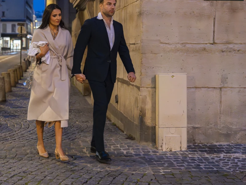 Jean-Yves · 15/05/2026 · 3000 x 2875 Cette photographie montre un couple marchant main dans la main dans une rue pavée. Le sujet principal est ce couple élégamment vêtu : elle porte une robe longue et légère, lui un costume sombre.La composition est symétrique et harmonieuse, avec un cadrage serré sur les deux personnages. L’arrière-plan, une pierre claire et un panneau indiquant "Place Saint-Sulpice", encadre la scène. La lumière douce, probablement artificielle, crée une ambiance tamisée et romantique. Les couleurs dominantes sont les tons neutres (beige, gris) avec des touches de noir et blanc, renforçant un style sophistiqué et intemporel. L’ambiance est calme et élégante.