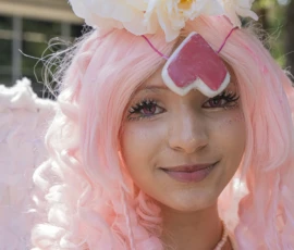 Jean-Yves · 24/05/2026 · 2261 x 3000 Cette photographie met en scène une personne au look fantaisiste. Le sujet principal est une jeune femme aux cheveux roses et bouclés, portant une coiffure décorée de fleurs blanches et d’un accessoire en forme de cœur rose.La composition est centrée sur son visage, avec un cadrage serré. La lumière est douce et naturelle, mettant en valeur les détails. Les couleurs dominantes sont le rose vif des cheveux et des accessoires, contrastant avec les tons pastel des fleurs.Le style est créatif et joyeux, évoquant une ambiance festive et décalée, typique d’un événement artistique ou d’une fête thématique.