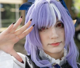 Jean-Yves · 24/05/2026 · 3000 x 3000 Cette photographie met en scène un individu déguisé en personnage stylisé, probablement inspiré d’un anime ou d’un manga. Le sujet principal est une personne avec une perruque violette à reflets blancs et des accessoires (ailes et maquillage). Le cadrage est serré, centré sur le visage et les épaules.La lumière est naturelle et douce, mettant en valeur les détails du maquillage et de la coiffure. Les couleurs dominantes sont le violet, le bleu et le noir, renforçant un style fantastique et cosplay. L’ambiance est festive et théâtrale, typique des conventions ou événements dédiés aux cultures otaku.