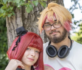 Jean-Yves · 24/05/2026 · 2845 x 3000 Cette photographie met en scène deux personnes déguisées en personnages stylisés, probablement inspirés de l’anime ou du manga. La jeune fille arbore une perruque rouge et un accessoire noir, tandis que l’homme porte une perruque blonde et des lunettes, avec des écouteurs.Le cadrage est serré, centré sur eux, avec un arbre en arrière-plan. La lumière naturelle est douce, éclairant leurs visages. Les couleurs dominantes sont vives : rouge, rose et jaune. Le style est décontracté et ludique, évoquant une ambiance festive, probablement liée à une convention ou un cosplay.