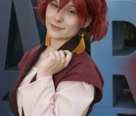 Jean-Yves · 24/05/2026 · 2000 x 3000 Cette photographie met en scène une personne déguisée en cosplay, probablement inspirée d'un personnage de manga ou d'anime. Le sujet principal est une femme aux cheveux roux, vêtue d'une tenue marron et blanche avec une cape.La composition est simple et centrée, avec un cadrage serré sur le personnage. L'arrière-plan montre une lettre géante "A" bleue, ajoutant un élément graphique distinctif. La lumière est naturelle et douce, éclairant bien les traits du visage. Les couleurs dominantes sont les tons chauds (rouge des cheveux, marron de la tenue) contrastant avec le bleu froid de l'arrière-plan. L'ambiance est festive et artistique, typique des conventions de cosplay.