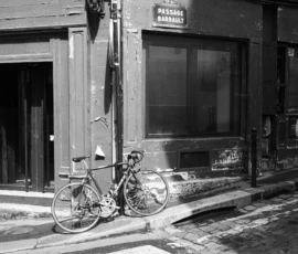 Agnès · 14/05/2026 · 6240 x 4160 Cette photographie en noir et blanc met en scène un vélo attaché à un réverbère devant une boutique urbaine. Le sujet principal est le vélo, simple et légèrement incliné, avec des détails comme les pneus et la selle bien visibles.La composition est épurée, centrée sur le vélo et le bâtiment en arrière-plan. Le cadrage est serré, capturant une partie de la rue et des éléments architecturaux.La lumière est douce, probablement naturelle, créant des contrastes modérés. Les couleurs dominantes sont les nuances de gris, typiques du noir et blanc.Le style est documentaire et intemporel, évoquant une ambiance calme et mélancolique, typique des scènes urbaines désertes.