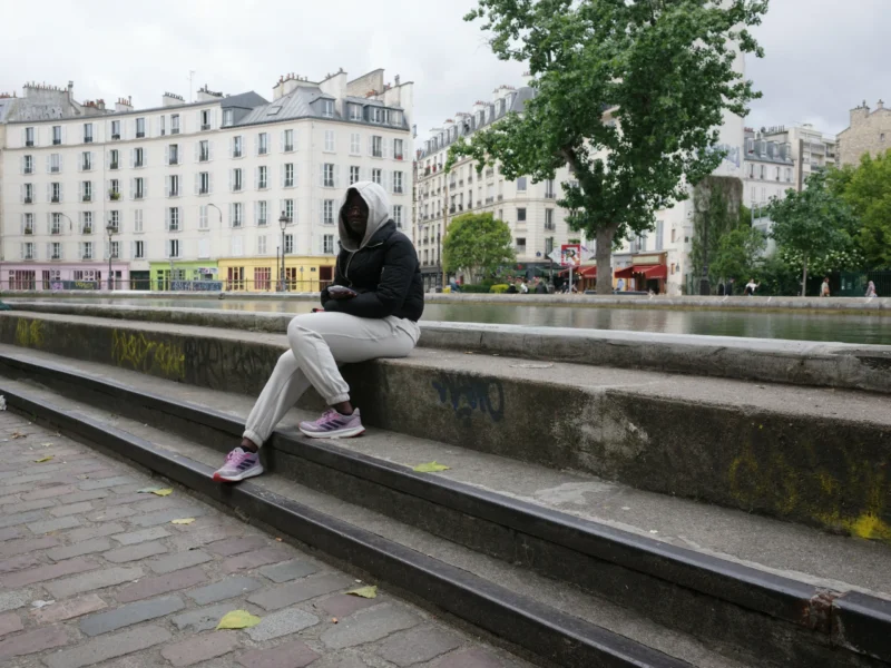 Bénédicte · 5504 x 4128 Cette photographie montre une personne assise sur une margelle de canal, le dos tourné à l'objectif. Le sujet principal est flou, ce qui met l'accent sur l'ambiance plutôt que sur les détails.La composition est équilibrée, avec le canal et les bâtiments en arrière-plan. Le cadrage est serré sur le sujet et son environnement immédiat. La lumière est naturelle, légèrement voilée par un ciel couvert, créant une ambiance douce et un peu mélancolique.Les couleurs dominantes sont les tons gris et beiges des bâtiments, avec des touches de vert des arbres et de rouge des éléments urbains. Le style est documentaire et réaliste, évoquant une scène de vie quotidienne.