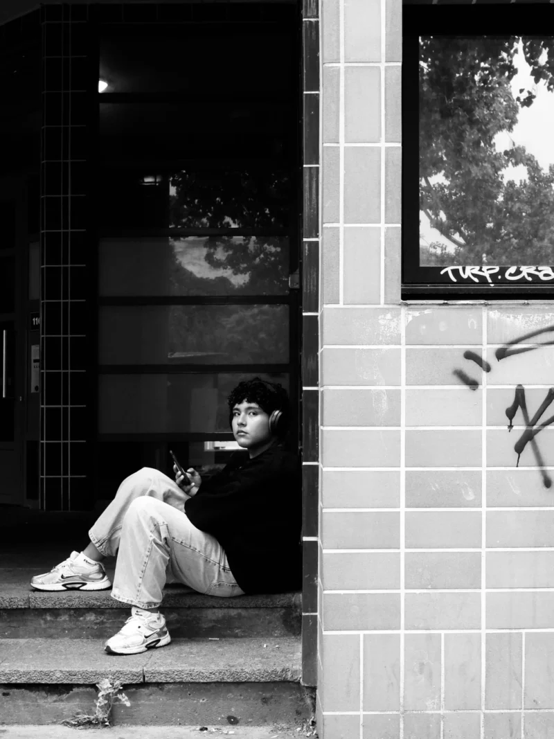 Bénédicte · 2305 x 3073 Cette photographie en noir et blanc met en scène un jeune homme assis sur des marches, près d’une porte entrouverte. Il semble absorbé par son téléphone portable.La composition est simple et équilibrée, avec un cadrage serré sur le sujet. La lumière est naturelle, douce mais contrastée, créant des ombres marquées. Les couleurs dominantes sont les tons de gris, renforçant une ambiance mélancolique et introspective.Le style rappelle le street photography, authentique et spontané. L’ambiance est calme et un peu solitaire, évoquant une pause dans le quotidien urbain.