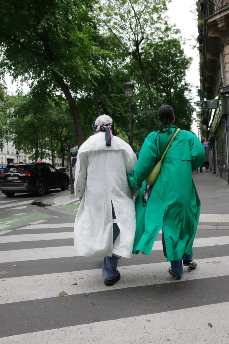 Bénédicte · 4128 x 6192 Cette photographie montre deux personnes marchant de dos sur un passage piéton. Le sujet principal est leur tenue colorée : l’un porte une longue robe blanche, l’autre une robe verte fluide. La composition est simple, axée sur leurs silhouettes en mouvement.Le cadrage est serré, centré sur les deux individus, avec un arrière-plan flou (voitures, arbres, bâtiments). La lumière semble naturelle, probablement en journée, avec des ombres douces. Les couleurs dominantes sont le blanc, le vert et les teintes grises urbaines. Le style est documentaire et spontané, créant une ambiance paisible et légèrement mystérieuse.