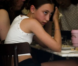 Pascale · 30/05/2026 · 963 x 1500 Cette photographie montre une jeune fille comme sujet principal, assise à une table, l’air pensif, la tête posée sur sa main. La composition est simple, avec un cadrage serré sur elle et une partie de l’espace autour.La lumière semble douce et naturelle, probablement artificielle, créant une ambiance tamisée. Les couleurs dominantes sont des tons chauds et neutres, avec des touches pastel sur les nappes et les vêtements. Le style est réaliste et spontané, évoquant une scène de vie quotidienne, peut-être dans un restaurant. L’ambiance est calme et introspective.