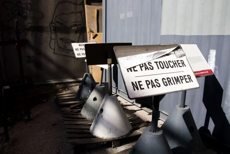 Pascale · 14/06/2026 · 1500 x 1008 Cette photographie montre un chantier avec des éléments métalliques (probablement des pièces industrielles) et une bâche partiellement visible. Le sujet principal est un panneau en français avertissant *« N'E PAS TOUCHER, NE PAS GRIMPER »*.La composition est simple, avec un cadrage serré sur les objets et le panneau. La lumière est naturelle, mais tamisée, créant des ombres marquées. Les couleurs dominantes sont les tons gris métalliques, noirs et blancs, avec des touches de rouge sur le panneau.Le style est documentaire, réaliste, évoquant une ambiance industrielle et sécuritaire, presque austère.