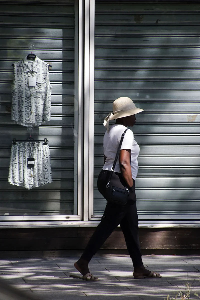 Pascale · 14/06/2026 · 1000 x 1500 Cette photographie met en scène une femme comme sujet principal, vêtue d’une tenue claire et d’un chapeau, marchant près d’un store métallique fermé. La composition est épurée, avec un cadrage serré qui isole la silhouette.La lumière, douce et contrastée, crée des ombres marquées, accentuant la texture des vêtements et du store. Les couleurs dominantes sont neutres, avec des tons gris et beiges, adoucis par des touches blanches.Le style est sobre et minimaliste, évoquant une ambiance calme et intemporelle, presque mélancolique. L’image suggère une scène urbaine paisible, imprégnée de sérénité.