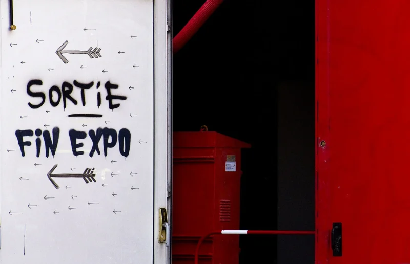 Pascale · 14/06/2026 · 1500 x 967 Cette photographie montre une porte avec un graffiti indiquant **« Sortie Fin Expo »** et des flèches directrices. Le sujet principal est ce message écrit à la peinture noire sur un fond blanc et grêlé.La composition est simple, avec un cadrage serré sur la porte rouge partiellement visible. La lumière est naturelle, contrastant les zones claires et sombres. Les couleurs dominantes sont le blanc, le noir et le rouge vif. Le style est brut et urbain, évoquant une ambiance d’abandon ou de fin d’événement, avec une touche d’art spontané.
