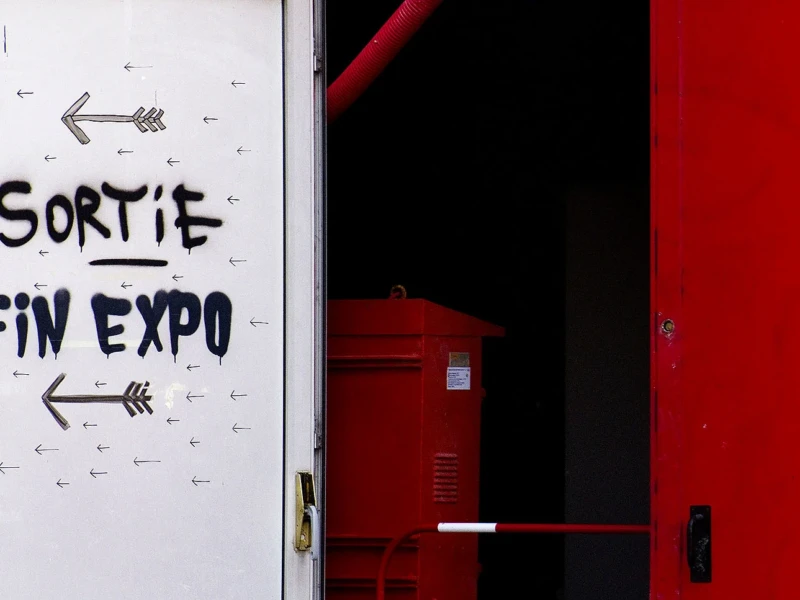 Pascale · 14/06/2026 · 1500 x 967 Cette photographie montre une porte avec un graffiti indiquant **« Sortie Fin Expo »** et des flèches directrices. Le sujet principal est ce message écrit à la peinture noire sur un fond blanc et grêlé.La composition est simple, avec un cadrage serré sur la porte rouge partiellement visible. La lumière est naturelle, contrastant les zones claires et sombres. Les couleurs dominantes sont le blanc, le noir et le rouge vif. Le style est brut et urbain, évoquant une ambiance d’abandon ou de fin d’événement, avec une touche d’art spontané.