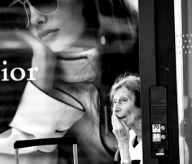 Franck T. · 907 x 1400 Cette photographie en noir et blanc met en scène une affiche publicitaire floue au premier plan, représentant une femme élégante avec des lunettes. En arrière-plan, une autre femme, partiellement visible, semble observer un écran de téléphone.La composition joue sur les reflets et les superpositions, créant une ambiance urbaine et moderne. Le cadrage est serré, centré sur les visages et les détails des vêtements. La lumière est douce, avec des contrastes marqués, accentuant les textures et les contours. Le style est épuré et contemporain, renforcé par le monochrome, évoquant une atmosphère à la fois dynamique et introspective.