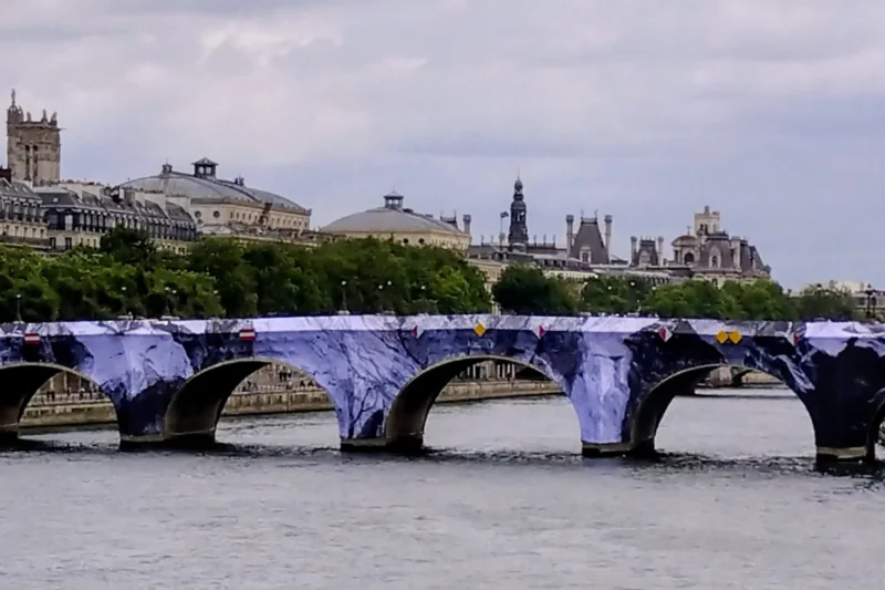Fabienne · 20/05/2026 · 1945 x 1297 Cette photographie met en scène un **pont couvert de graffitis violets** en premier plan, traversant un plan d’eau. Le **sujet principal** est ce pont, dont les arches sont soulignées par les couleurs vives.La **composition** est équilibrée, avec une perspective légèrement basse, renforçant la profondeur. Le **cadrage** inclut des bâtiments et une colline en arrière-plan, ajoutant de la dimension.La **lumière** est naturelle, douce, sans ombre marquée. Les **couleurs dominantes** sont les tons violets du graffiti, contrastant avec le bleu du ciel et de l’eau.Le style est **urbain et artistique**, évoquant une ambiance à la fois **moderne et mélancolique**.