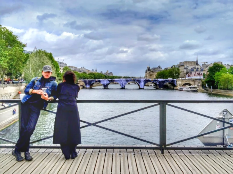 Fabienne · 20/05/2026 · 2885 x 2164 Cette photographie montre deux personnes debout sur un pont en bois, le long d'une rivière. Le sujet principal semble être leur interaction, avec une ambiance intime et contemplative.La composition est équilibrée, avec le pont en premier plan et les ponts en arrière-plan. Le cadrage inclut une vue large de la rivière et des bâtiments.La lumière est naturelle et douce, typique d'une journée légèrement nuageuse. Les couleurs dominantes sont les tons bleus de l’eau et du ciel, avec des touches de gris et de vert.Le style est réaliste et photographique, évoquant une ambiance paisible et romantique.