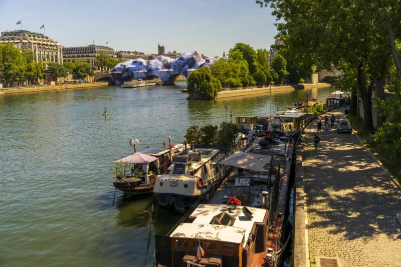 Thierry · 22/05/2026 · 3840 x 2560 Cette photographie capture une scène fluviale animée. Le **sujet principal** est un **bateau-hôtel** amarré le long des berges, avec des passagers détendus.La **composition** alterne entre le **cours d’eau**, les **bateaux** et les **passants** sur les quais. Le **cadrage** est large, incluant aussi des **arbres**, des **bâtiments** et des **installations artistiques** (sculptures bleues).La **lumière** est naturelle et généreuse, créant des ombres douces. Les **couleurs dominantes** sont les **tons bleus** (eau, ciel), **verts** (végétation) et **beiges/gris** (bâtiments, bateaux).Le **style** est réaliste et documentaire, évoquant une **ambiance paisible et estivale**.