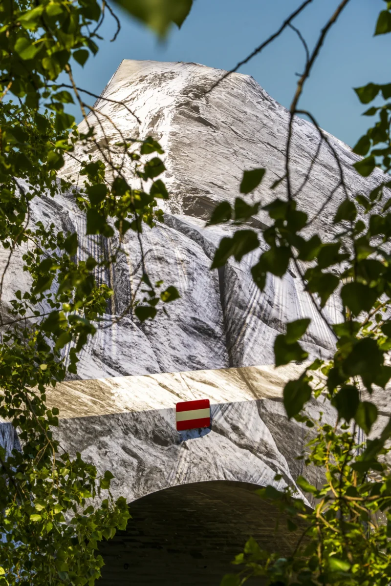 Thierry · 22/05/2026 · 2560 x 3840 Cette photographie met en scène une grande tête sculptée dans la roche, partiellement cachée par des feuilles vertes. Le sujet principal est cette tête, dont les traits sont stylisés et imposants.La composition joue sur le cadrage serré, encadrant la tête entre les branches. La lumière naturelle, vive et contrastée, accentue les textures de la pierre et les ombres. Les couleurs dominantes sont les gris de la roche, les verts des feuilles et le rouge du panneau en bas à gauche.L’ambiance est mystérieuse et monumentale, évoquant une œuvre artistique ou un site historique. Le style semble moderne et épuré.