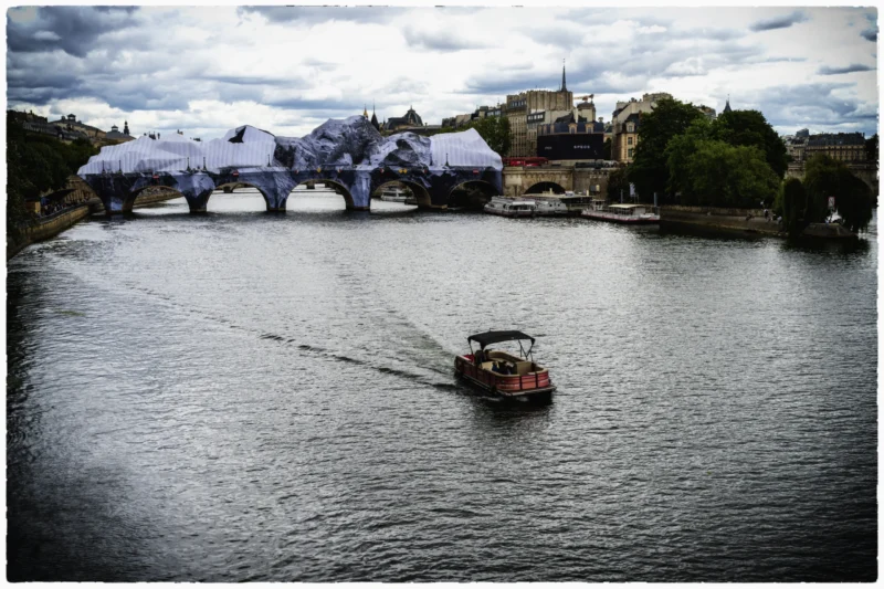 Thierry · 11/06/2026 · 3840 x 2560 Cette photographie met en scène un **bateau rouge** comme sujet principal, naviguant sur une rivière. La **composition** est équilibrée, avec le bateau centré et un **pont couvert** en arrière-plan, partiellement voilé de bâches blanches.Le **cadrage** est large, capturant aussi des bâtiments et des arbres. La **lumière** est tamisée, créant une ambiance sobre et légèrement mélancolique. Les **couleurs dominantes** sont les tons gris-bleutés de l’eau et du ciel, contrastant avec le rouge vif du bateau.Le style est **réaliste et contemplatif**, évoquant une atmosphère calme et presque nostalgique.