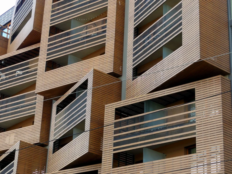 Marine · 14/06/2026 · 4000 x 3000 Cette photographie montre la façade d’un immeuble moderne. Le sujet principal est l’architecture géométrique et les balcons en bois disposés en angles variés. La composition met en valeur les lignes dynamiques et les jeux d’ombres et de lumières.Le cadrage est serré, centré sur les balcons. La lumière naturelle éclaire les surfaces, créant des contrastes nets. Les couleurs dominantes sont les tons bois naturels et les teintes grises des structures métalliques. Le style est contemporain et épuré, évoquant une ambiance à la fois minimaliste et innovante.
